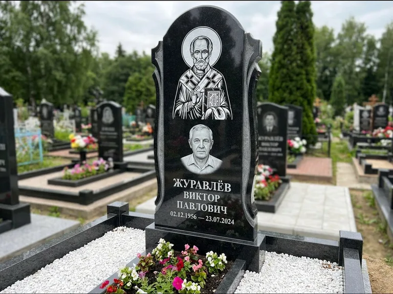 Памятник с гравировкой иконы: стела с образом в полный рост на обороте — гравировка образа Матроны Московской, низкие кусты спиреи в цвету, работа «Лебедева» в Ростове-на-Дону
